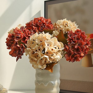 May include: A bouquet of artificial hydrangea flowers in a white ceramic vase. The flowers are in shades of red, brown, and cream.
