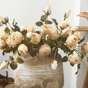 Könnte beinhalten: Ein Strauß künstlicher Pfirsichrosen in einer cremefarbenen Keramikvase mit strukturierter Oberfläche. Die Rosen sind in einem lockeren, natürlichen Stil angeordnet.