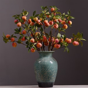 Peut inclure: Un vase en céramique vert avec un motif moucheté contient un bouquet de branches de pêche artificielles avec des fruits rouges et orange.