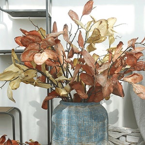 May include: A display of artificial autumn leaves in a textured blue vase. The arrangement includes branches with large, dried-looking leaves in shades of orange, brown, and yellow. The vase has a rustic, weathered appearance, enhancing the natural aesthetic.