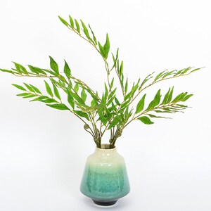 Può includere: Un vaso in ceramica verde con una glassa testurizzata contiene un bouquet di rami verdi con foglie.