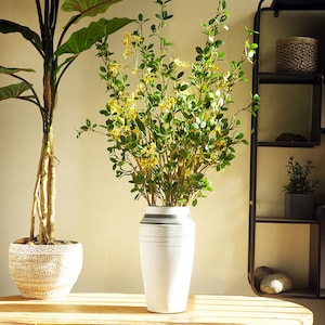 May include: A white ceramic vase with gray stripes holds a bouquet of artificial green leaves with yellow flowers. The vase is sitting on a wooden table.