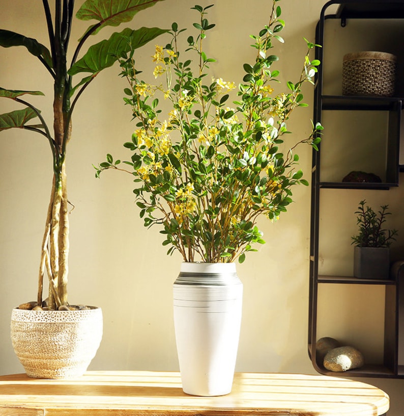 May include: A white ceramic vase with gray stripes holds a bouquet of artificial green leaves with yellow flowers. The vase is sitting on a wooden table.