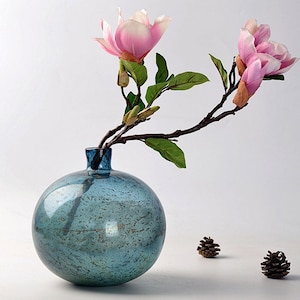 Könnte beinhalten: Eine blaue Glasvase mit strukturierter Oberfläche, die einen Zweig mit rosa Magnolienblüten hält.