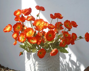 Puede incluir: Un ramo de flores artificiales de amapola de color naranja y amarillo brillante en un jarrón blanco con un patrón texturizado.