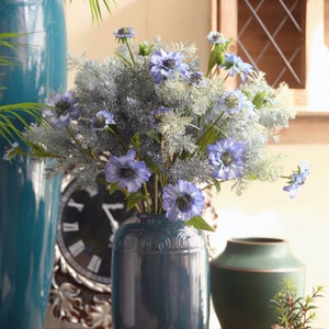 May include: A bouquet of blue and white artificial flowers in a dark teal vase. The vase has a decorative design and is sitting on a wooden table.
