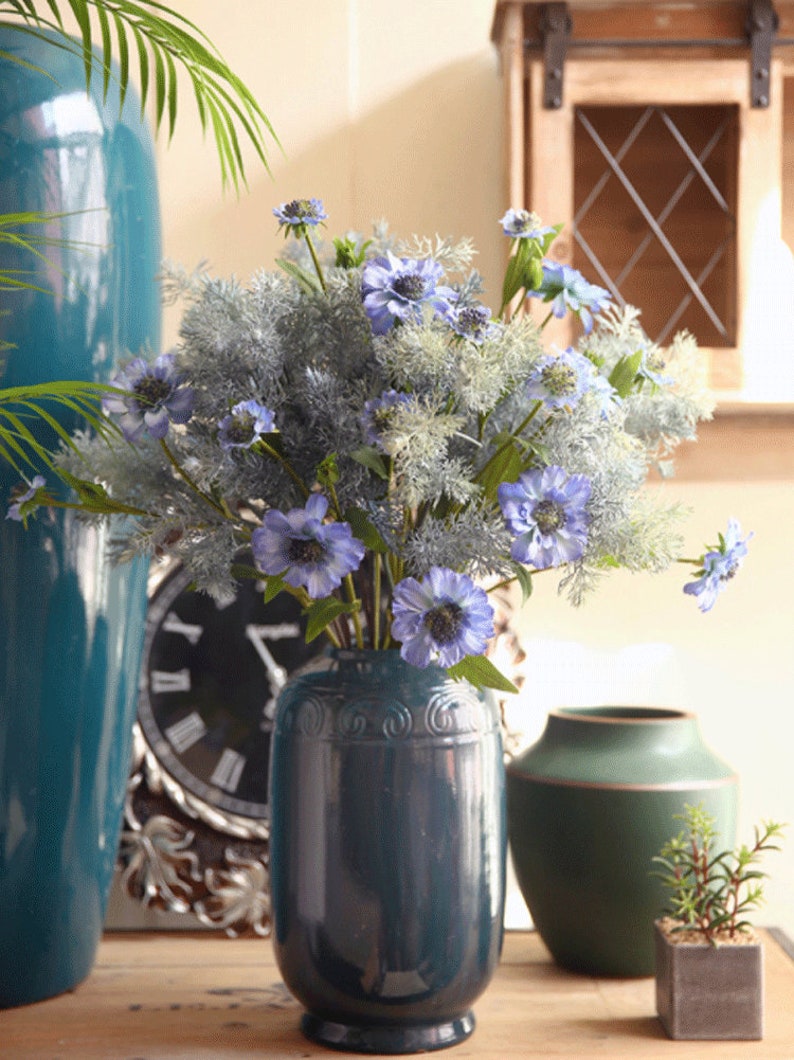May include: A bouquet of blue and white artificial flowers in a dark teal vase. The vase has a decorative design and is sitting on a wooden table.