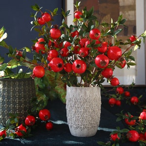 May include: A bouquet of artificial pomegranate branches with red fruit and green leaves in a grey ceramic vase with a geometric pattern. The vase is sitting on a dark blue surface.
