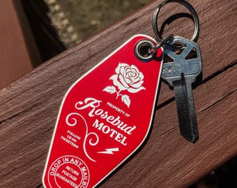 May include: A red motel key tag with the words "Rosebud Motel" and a white rose graphic. A silver key is attached to the tag with a metal ring. The tag also has the text "Drop in any mailbox, return postage guaranteed."