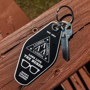 May include: Black key tag with a silver key attached. The tag features white text and graphics, including the words "Lambda," "Long Live The Nerds," and "Adam's College Alumni." The tag is on a wooden surface.