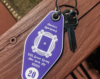 May include: A purple key tag with the text "Monica's Apartment" and the address "545 Grove Street New York, NY 10001". The tag also features the number "20" and a key ring with a key.
