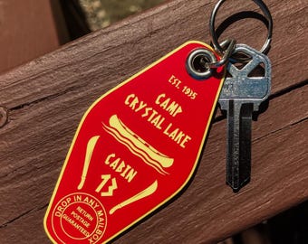May include: A red motel-style keychain with the text "Camp Crystal Lake Cabin 13" and a key. The keychain has yellow accents and the words "Drop in any mailbox" and "Return postage guaranteed".