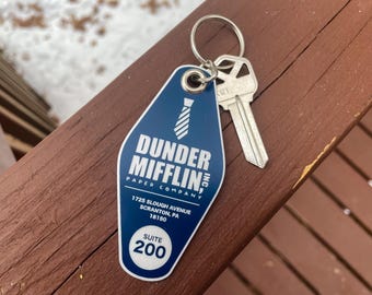 May include: A blue Dunder Mifflin Paper Company keychain with a silver key attached. The keychain has the company logo, address, and suite number printed on it. The keychain is on a brown wooden surface.