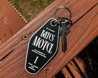 May include: A black and white Bates Motel key tag with a silver key attached. The tag reads "NO VACANCY" and "BATES MOTEL" with the address "OLD HIGHWAY 10 FAIRVALE, CALIFORNIA" and "1". The tag also says "FAMILY OWNED AND OPERATED".