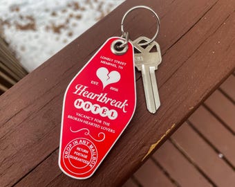 May include: A red Heartbreak Hotel keychain with a silver key. The keychain has the text "Heartbreak Hotel" and "Vacancy for the broken-hearted lovers." The keychain also has a broken heart graphic. The key is silver.
