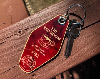 May include: A vintage motel key tag with a red background and the words "THE RED ROOM" and "HILL HOUSE MASSACHUSETTS 1992". The key tag has a silver key ring and a silver key attached. The key tag is on a wooden surface.