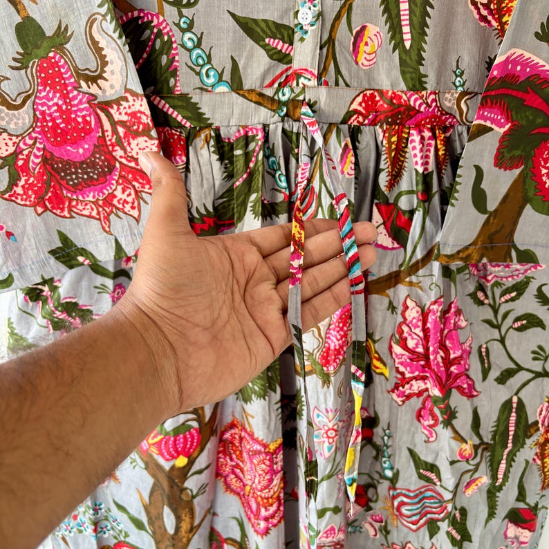 May include: A gray cotton dress with a floral print featuring pink, green, and blue flowers. The dress has a drawstring waist and long sleeves.
