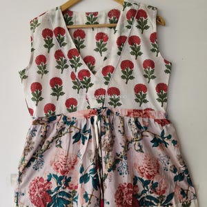 Puede incluir: Un vestido sin mangas con escote en V y cintura con cordón ajustable. El corpiño tiene un fondo blanco con un estampado floral rojo y verde. La sección de la falda muestra un patrón floral más grande en rosa, rojo y verde sobre fondo claro.