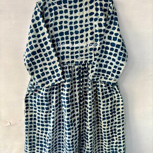 Puede incluir: Vestido de manga larga con fondo blanco y un patr&oacute;n repetitivo de cuadrados azul oscuro. El vestido tiene cuello redondo, cintura fruncida y bolsillos. El vestido est&aacute; colgado de una percha de madera.