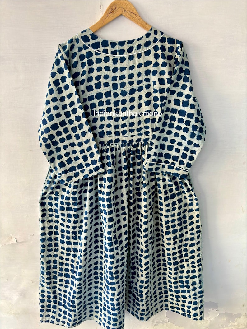 Puede incluir: Vestido de manga larga con fondo blanco y un patr&oacute;n repetitivo de cuadrados azul oscuro. El vestido tiene cuello redondo, cintura fruncida y bolsillos. El vestido est&aacute; colgado de una percha de madera.