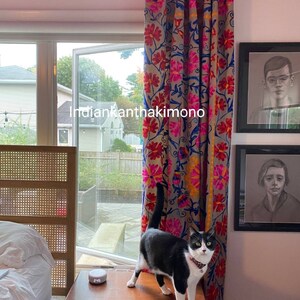 Puede incluir: Un gato blanco y negro se sienta en una mesa de madera frente a una ventana con vista a un patio trasero. El gato mira hacia la derecha de la imagen. La ventana est&aacute; cubierta con una cortina de estampado floral colorido. El texto "indiankantha kimono" es visible en la cortina.