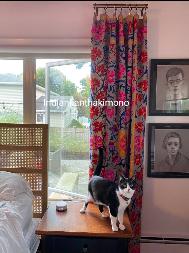 Puede incluir: Un gato blanco y negro se sienta en una mesa de madera frente a una ventana con vista a un patio trasero. El gato mira hacia la derecha de la imagen. La ventana est&aacute; cubierta con una cortina de estampado floral colorido. El texto "indiankantha kimono" es visible en la cortina.