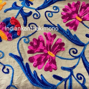 Puede incluir: Primer plano de una tela beige con bordados coloridos. El bordado presenta flores rosas, azules y naranjas con detalles intrincados. El texto "indiankanthakimono" es visible en el centro de la imagen.