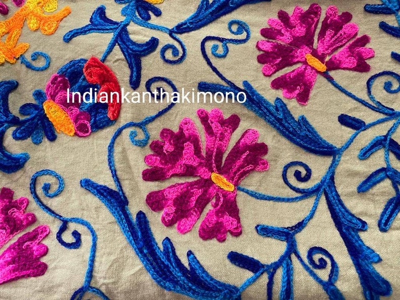 Puede incluir: Primer plano de una tela beige con bordados coloridos. El bordado presenta flores rosas, azules y naranjas con detalles intrincados. El texto "indiankanthakimono" es visible en el centro de la imagen.