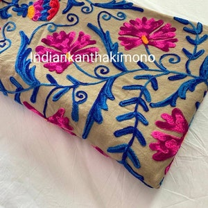 Puede incluir: Tela beige con bordado floral de colores. El bordado presenta flores rosas, azules y naranjas con hojas verdes. La tela est&aacute; doblada por la mitad y el texto "indiankantha kimono" es visible.