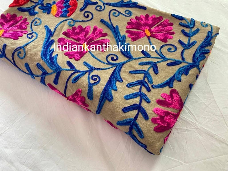 Puede incluir: Tela beige con bordado floral de colores. El bordado presenta flores rosas, azules y naranjas con hojas verdes. La tela est&aacute; doblada por la mitad y el texto "indiankantha kimono" es visible.