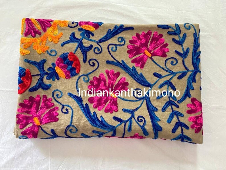 Puede incluir: Tela beige con bordado floral de colores. El bordado presenta flores rosas, azules, naranjas y amarillas. El texto "Indiankanthakkimono" es visible en el centro de la tela.