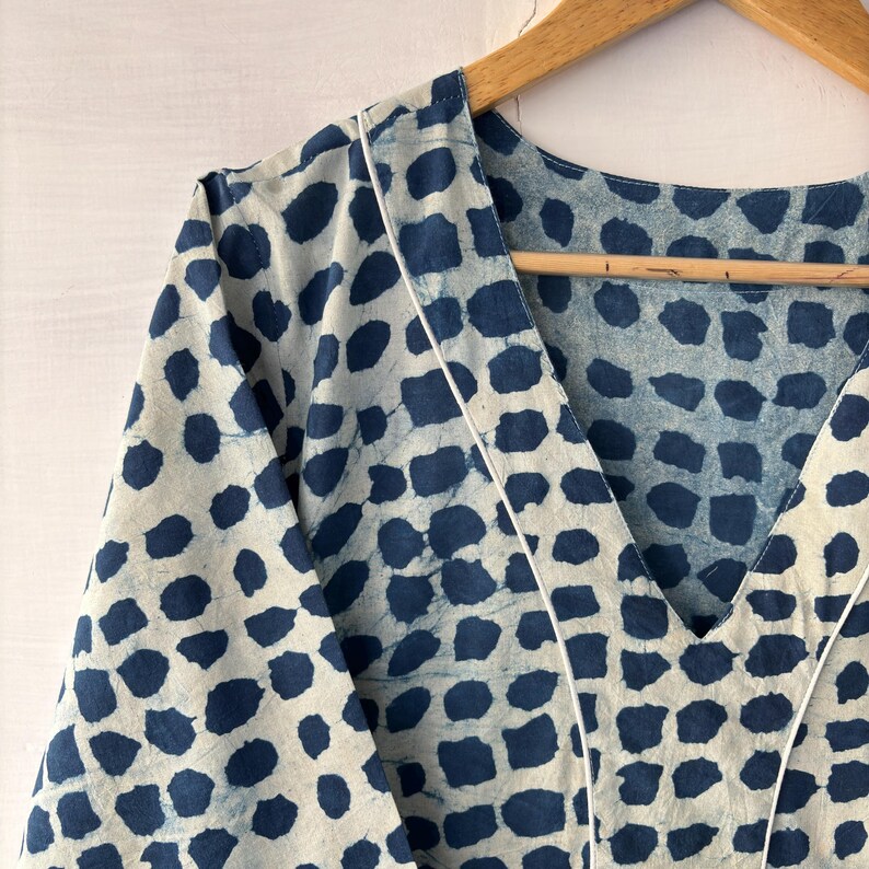 Puede incluir: Prenda de color claro con cuello en V y un patr&oacute;n repetitivo de formas irregulares de color azul oscuro. Los bordes de la prenda est&aacute;n ribeteados con un fino borde blanco. La tela est&aacute; colgada de una percha de madera.