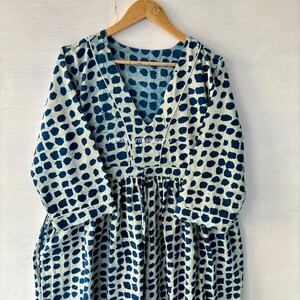 Puede incluir: Vestido de manga larga con cuello en V y cintura fruncida. El vestido presenta un estampado de puntos azul oscuro sobre un fondo crema. Ribetes blancos delinean el escote y las mangas. El vestido est&aacute; colgado de una percha de madera.