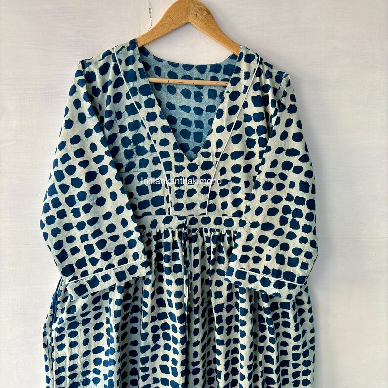Puede incluir: Vestido de manga larga con cuello en V y cintura fruncida. El vestido presenta un estampado de puntos azul oscuro sobre un fondo crema. Ribetes blancos delinean el escote y las mangas. El vestido est&aacute; colgado de una percha de madera.
