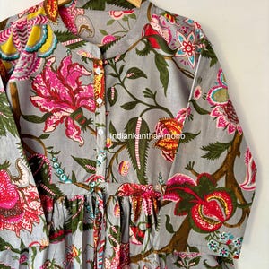May include: A gray cotton tunic with a floral print featuring pink, red, yellow, and blue flowers. The tunic has a button-up front and a tie at the waist.