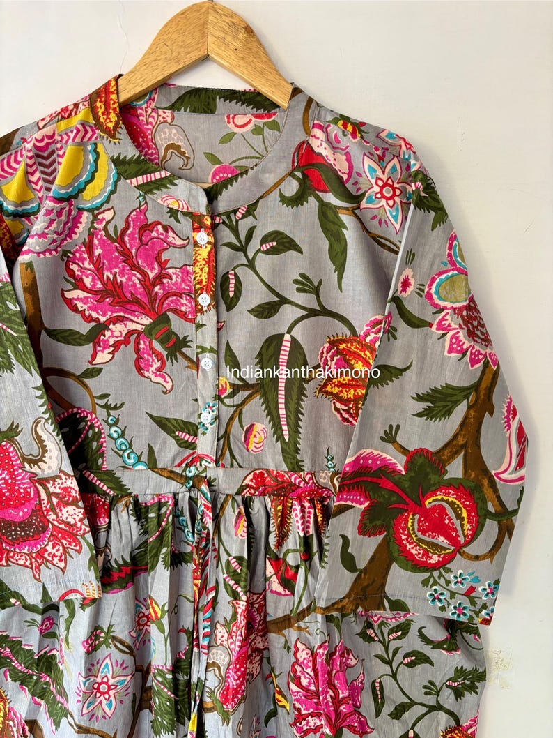 May include: A gray cotton tunic with a floral print featuring pink, red, yellow, and blue flowers. The tunic has a button-up front and a tie at the waist.