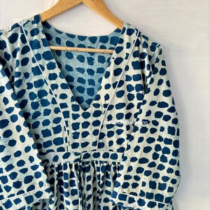 Puede incluir: Un vestido color crema con un estampado de lunares azules. El vestido tiene un escote en V, mangas largas y cintura fruncida. Ribetes blancos delinean el escote y las mangas. El vestido est&aacute; colgado de una percha de madera.