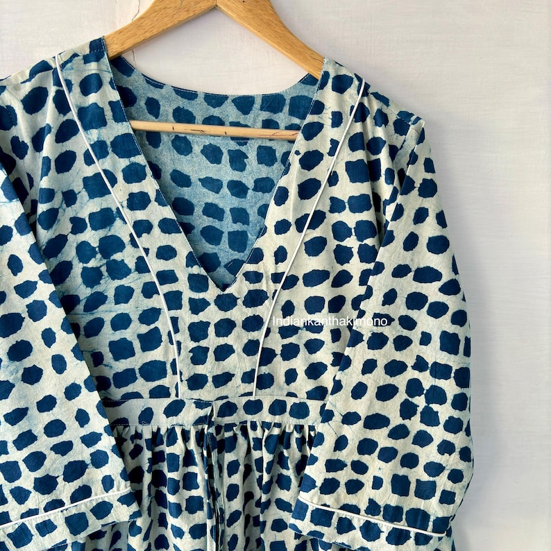 Puede incluir: Un vestido color crema con un estampado de lunares azules. El vestido tiene un escote en V, mangas largas y cintura fruncida. Ribetes blancos delinean el escote y las mangas. El vestido est&aacute; colgado de una percha de madera.
