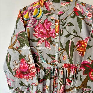 May include: A gray cotton tunic with a floral print featuring pink, red, yellow, and green flowers. The tunic has a mandarin collar and long sleeves. The fabric is gathered at the waist.