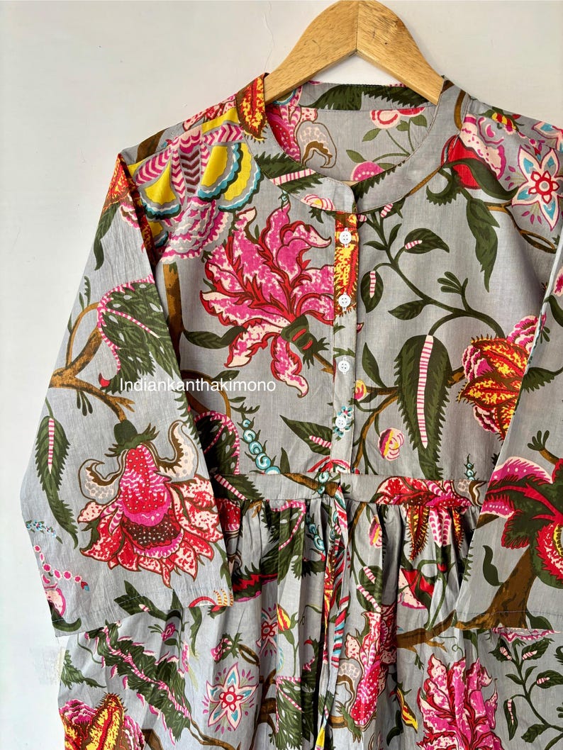 May include: A gray cotton tunic with a floral print featuring pink, red, yellow, and green flowers. The tunic has a mandarin collar and long sleeves. The fabric is gathered at the waist.