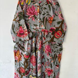 May include: A gray cotton dress with a floral print. The dress has a round neckline and long sleeves. The dress has a button closure at the neckline. The dress has a gathered skirt. The dress has a floral print with pink, red, yellow, and green flowers. The dress has the text "Tapestryhubindia" printed on the front.