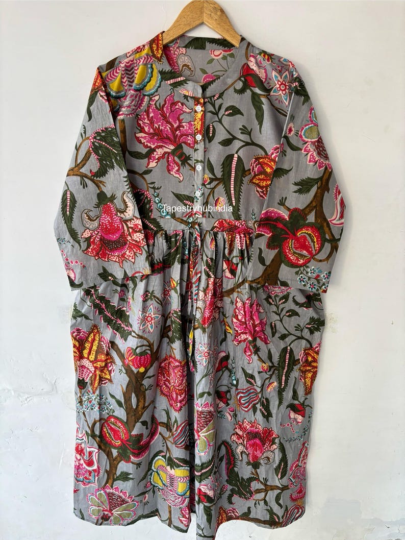 May include: A gray cotton dress with a floral print. The dress has a round neckline and long sleeves. The dress has a button closure at the neckline. The dress has a gathered skirt. The dress has a floral print with pink, red, yellow, and green flowers. The dress has the text "Tapestryhubindia" printed on the front.