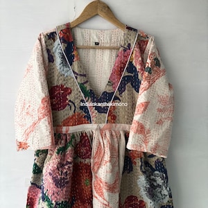 May include: A white cotton kantha dress with a floral print and a V-neckline. The dress has a tie at the waist and 3/4 sleeves. The dress is made in India.