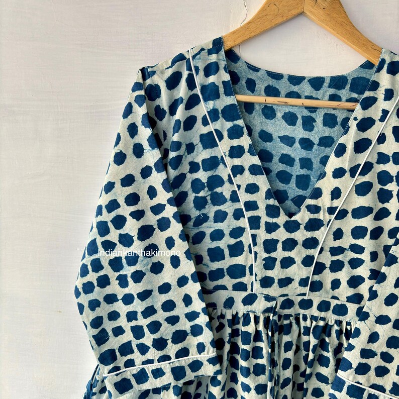 Puede incluir: Vestido de manga larga con cuello en V en un patr&oacute;n azul claro y blanco. El vestido presenta un patr&oacute;n repetitivo de cuadrados azul oscuro. Ribetes blancos delinean el escote y las mangas. El vestido est&aacute; colgado en una percha de madera.