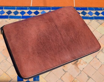 May include: A brown leather laptop sleeve with a zipper closure. The sleeve is lying on a tiled surface.