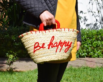 Puede incluir: Un bolso de mimbre con asas rojas y bordado rojo que dice "Be happy".