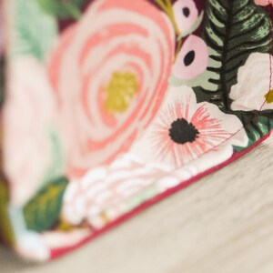 Pu&ograve; includere: Primo piano di un tessuto color borgogna con una stampa floreale con fiori rosa, bianchi e verdi. Il tessuto ha una cucitura rossa lungo il bordo.