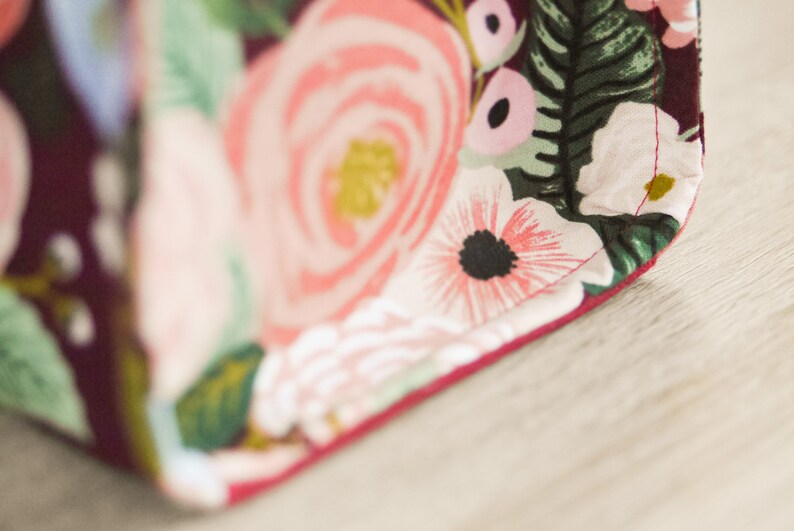 Pu&ograve; includere: Primo piano di un tessuto color borgogna con una stampa floreale con fiori rosa, bianchi e verdi. Il tessuto ha una cucitura rossa lungo il bordo.