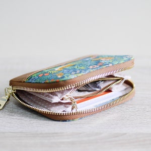 May include: A small, floral patterned zippered wallet with a brown leather trim. The wallet is open and shows a pink and white patterned lining, a few cards, and some coins.