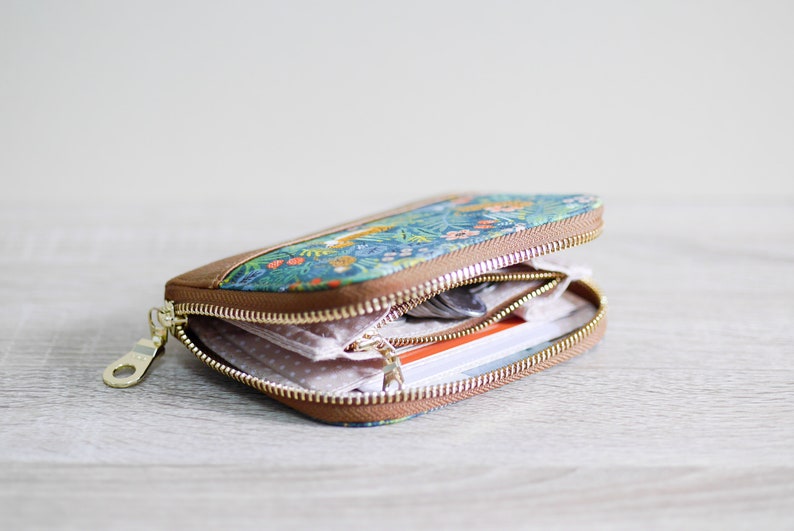 May include: A small, floral patterned zippered wallet with a brown leather trim. The wallet is open and shows a pink and white patterned lining, a few cards, and some coins.